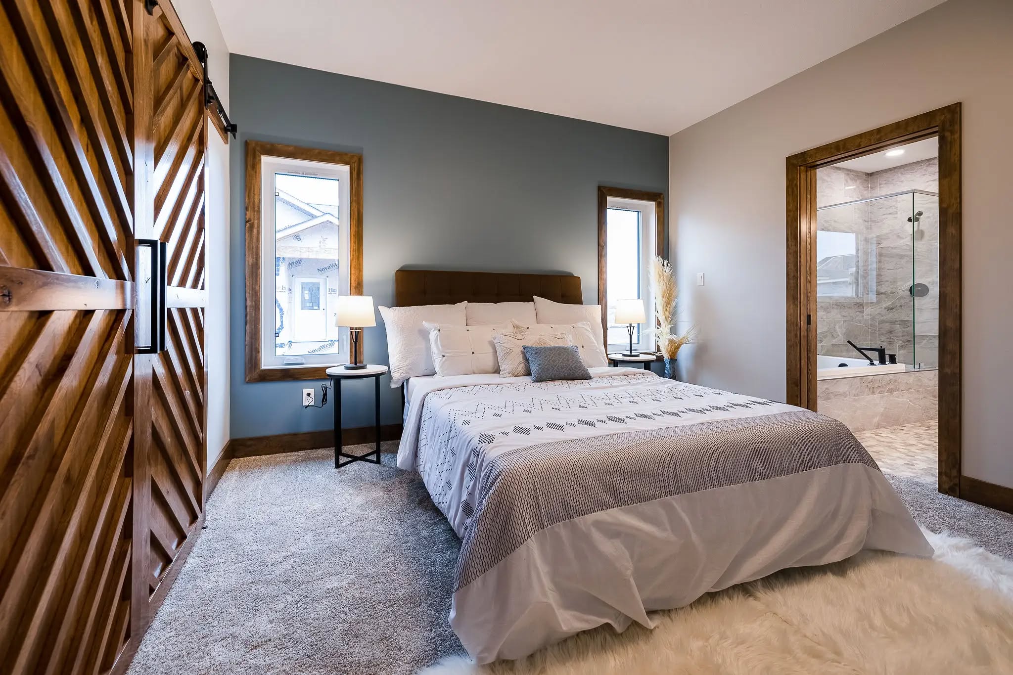 Cozy bedroom with a beige and gray color scheme, featuring a bed, a nightstand with a lamp, wooden sliding door, and an open ensuite bathroom.