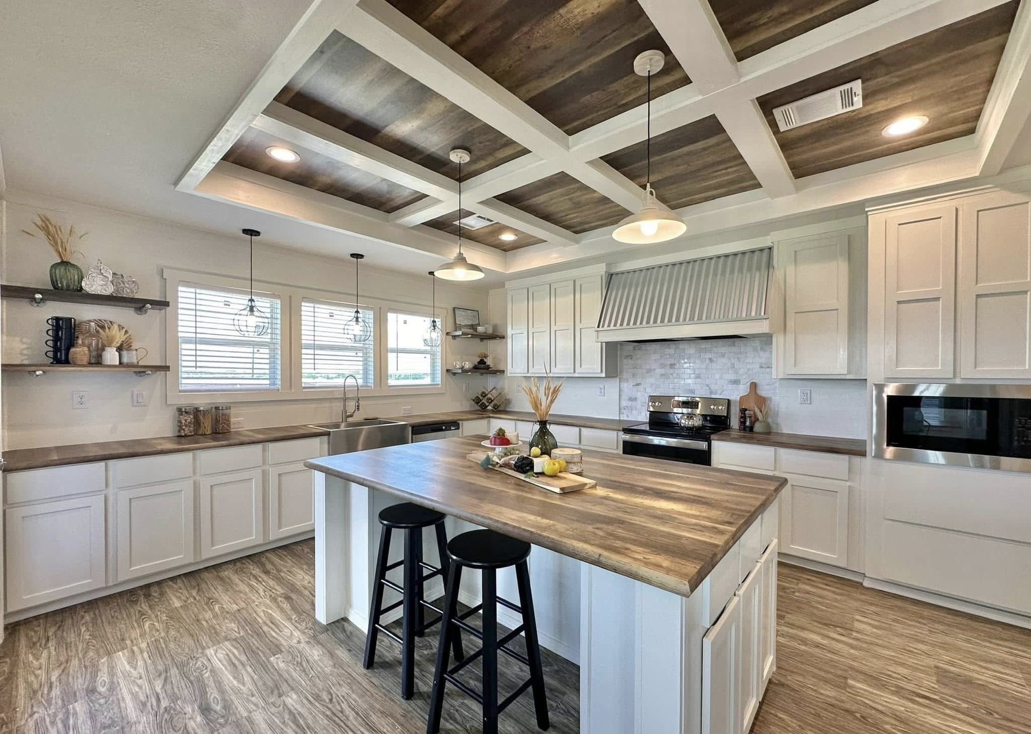 Spacious kitchen with white cabinets, wooden island, and two black stools. Pendant lights hang from a coffered ceiling. Natural light from large windows.