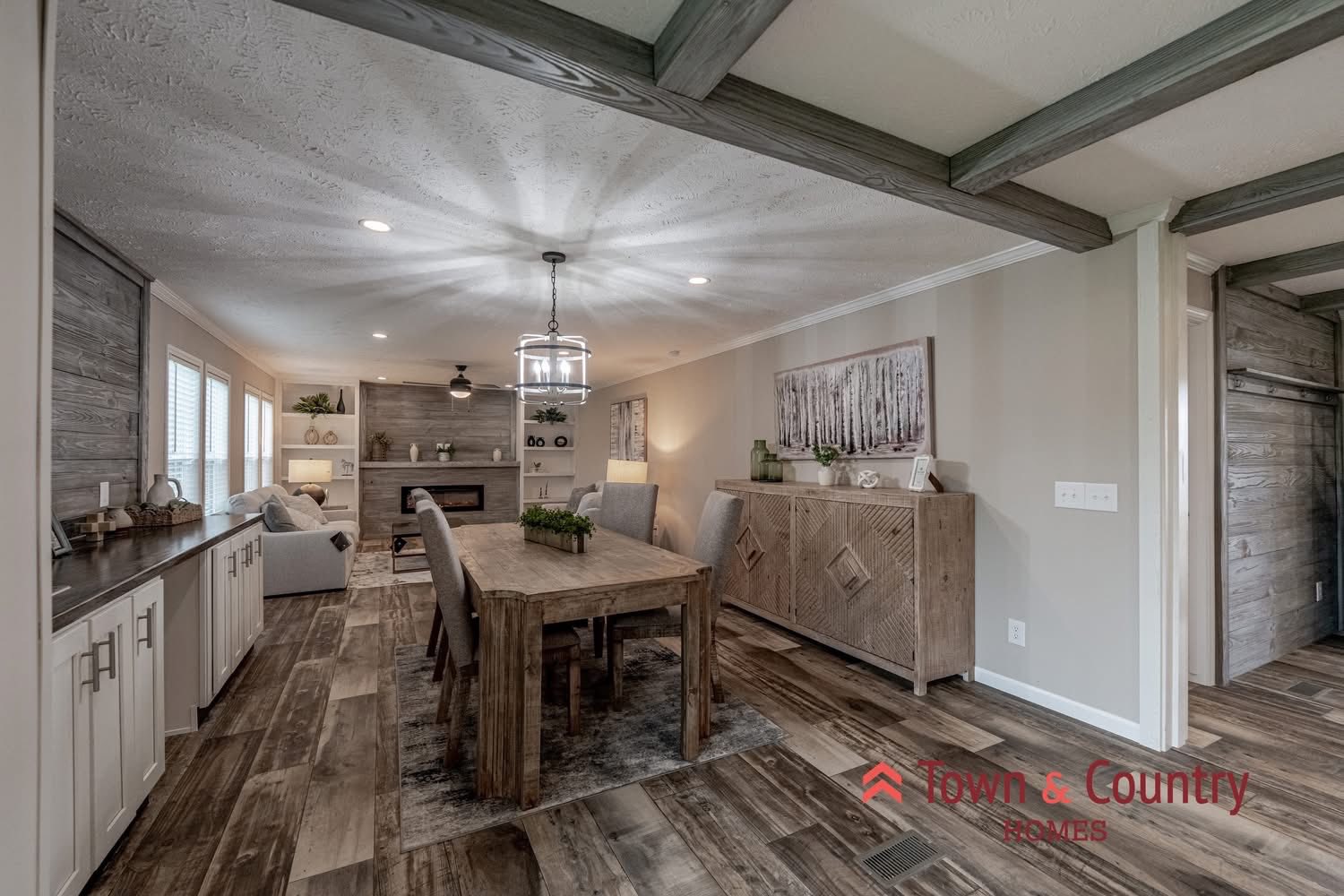 Modern open-concept living room and dining area featuring wooden floors, a rustic dining table, and cozy seating around a fireplace, conveying warmth and elegance.