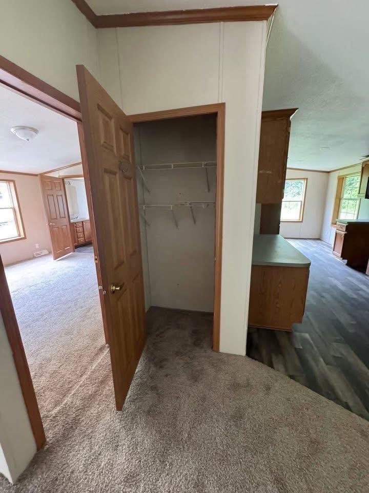 Open closet with wooden door in a carpeted room, adjacent to a room with wooden flooring and cabinets. Bright natural light from windows. Cozy and inviting.