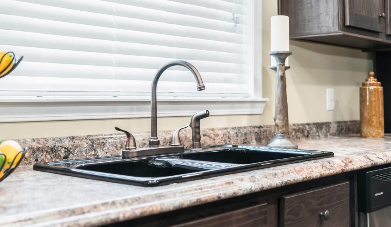 A modern kitchen sink with a high-arching faucet on a marbled countertop. It’s flanked by a lit candle and decorative vase, under a window with blinds.