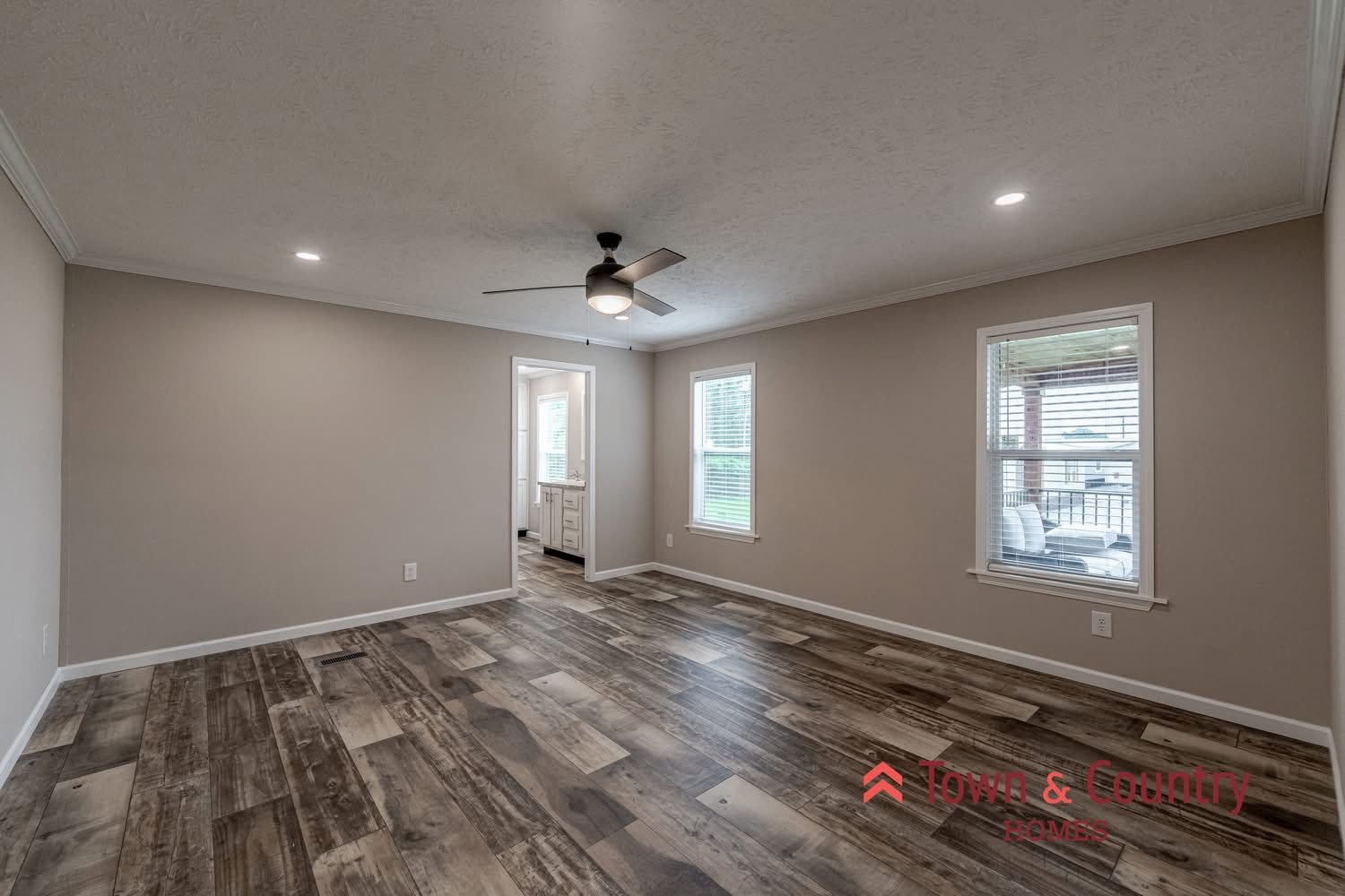 A spacious, empty room with wood-look flooring and neutral walls, featuring recessed lighting and a ceiling fan. Two windows provide natural light.