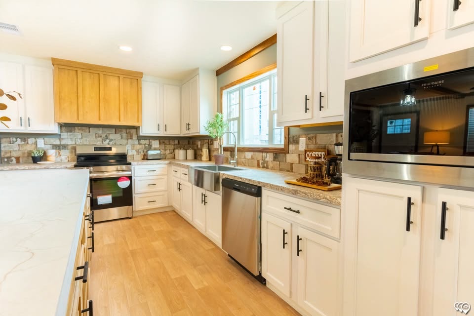 Modern kitchen with wooden floors, white cabinets, and stainless steel appliances. Stone backsplash, a farmhouse sink, and a potted plant by the window.