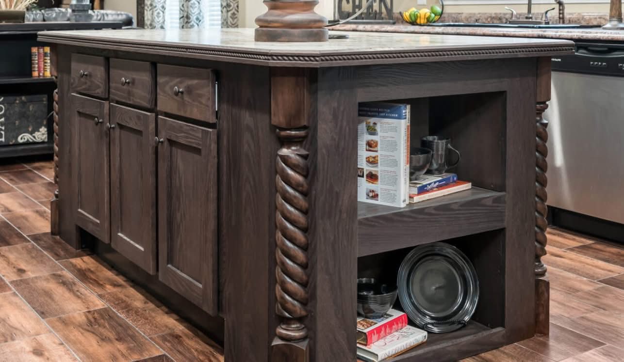 Rustic kitchen island with dark wood finish, ornate rope-like carvings, and a marble top. Open shelves hold books and dishes, evoking a warm, homey feel.