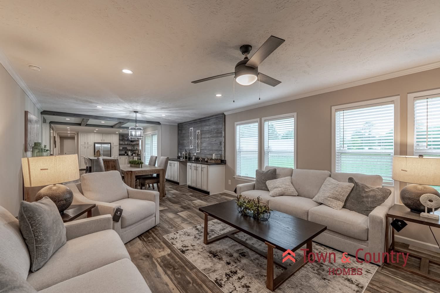 Spacious living room with neutral tones, cozy sofas, and a coffee table. Modern ceiling fan, natural light from large windows; open kitchen in the background.