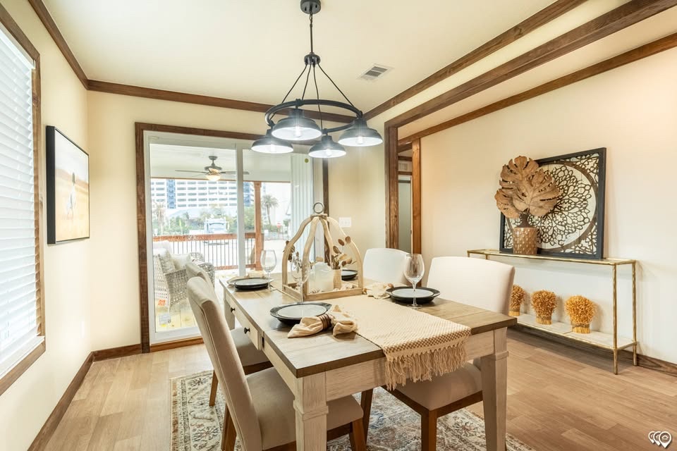 A cozy dining room with a light wooden table set for four, featuring black plates and beige napkins. Elegant lighting hangs above. A decorative console with art and dried plants adorns the back, while glass doors reveal a balcony view.
