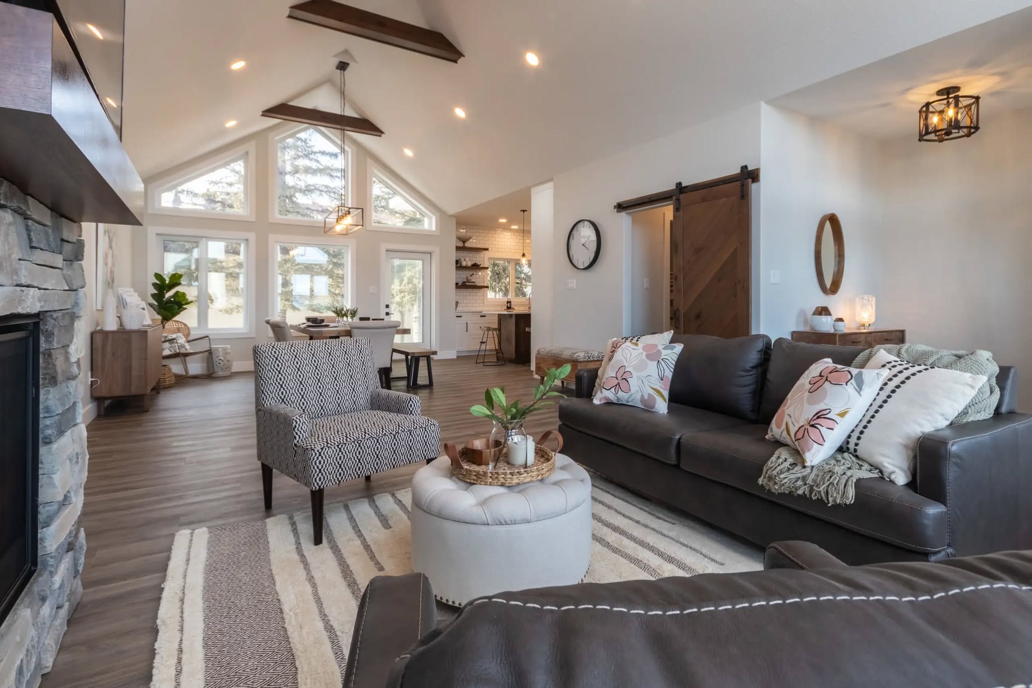Spacious living room with plush sofas, patterned armchair, stone fireplace, vaulted ceilings, large windows, and rustic decor, exuding warmth.