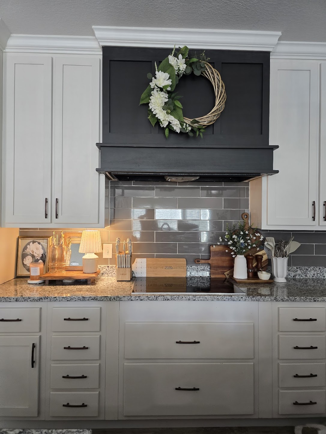 Elegant kitchen with white cabinets, granite countertops, and a gray tile backsplash. A floral wreath adorns the black range hood, adding charm.