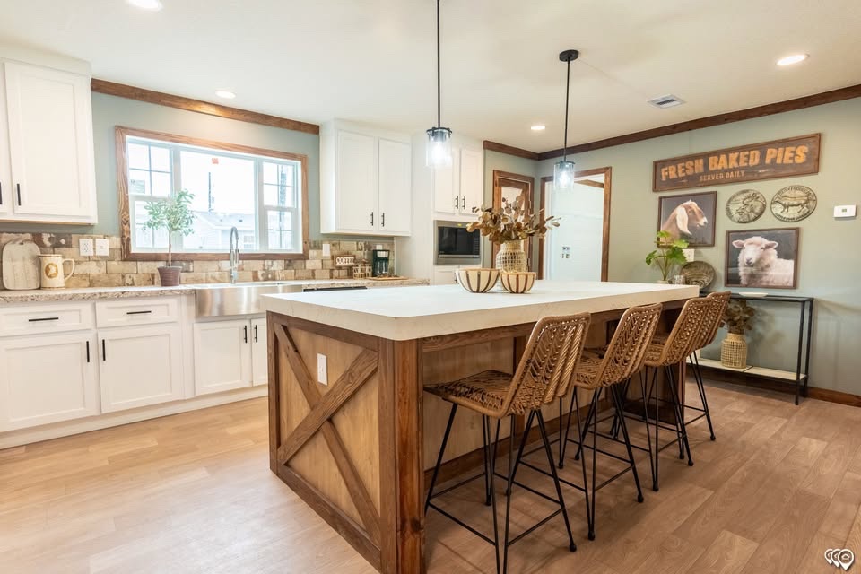 A rustic kitchen with a large wooden island, wicker chairs, and hanging lights. White cabinets and farmhouse decor create a cozy, inviting atmosphere.