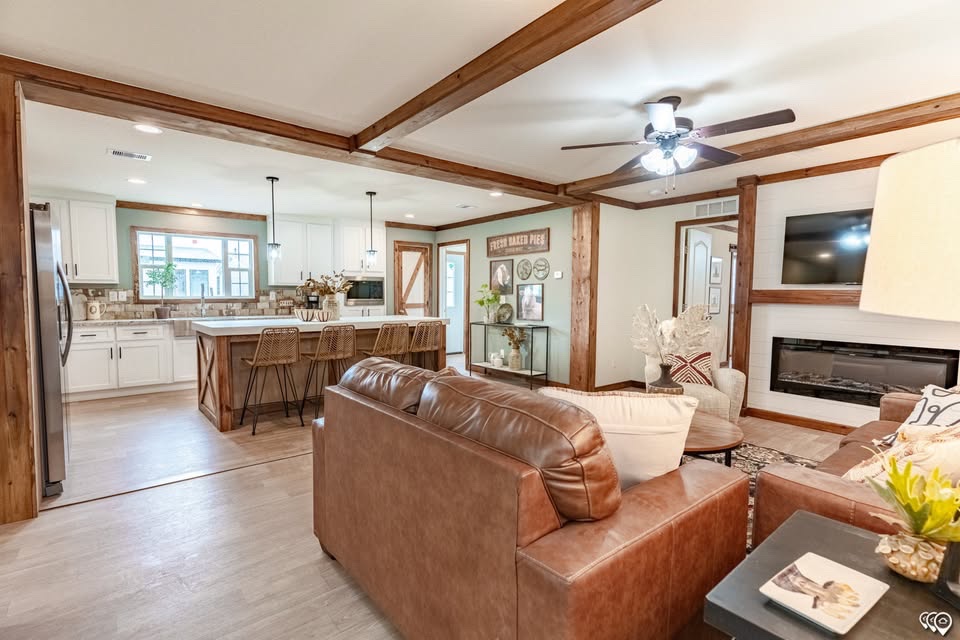 Cozy living room with brown leather sofas, ceiling fan, and fireplace. Open layout leads to modern kitchen with island stools and farmhouse decor.