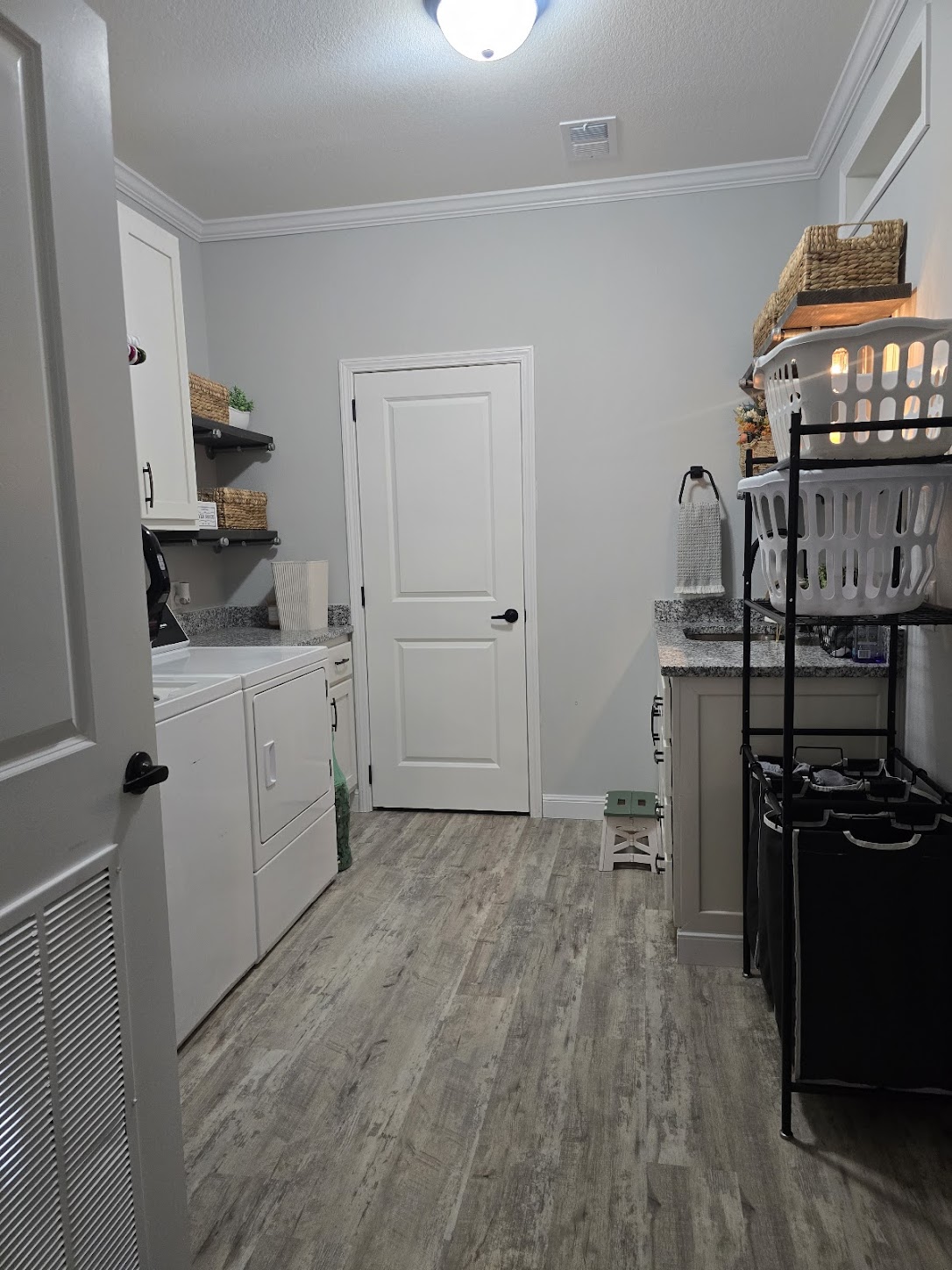 Laundry room with light wood flooring, a white washer and dryer on the left, a countertop with shelves and baskets on the right, and a white door.