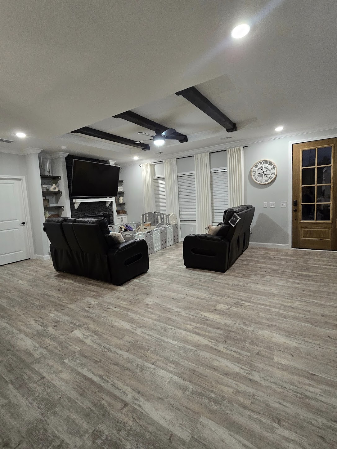 A cozy living room features two black recliners facing a wall-mounted TV above a stone fireplace. Light wood flooring, ceiling beams, and soft lighting add warmth.