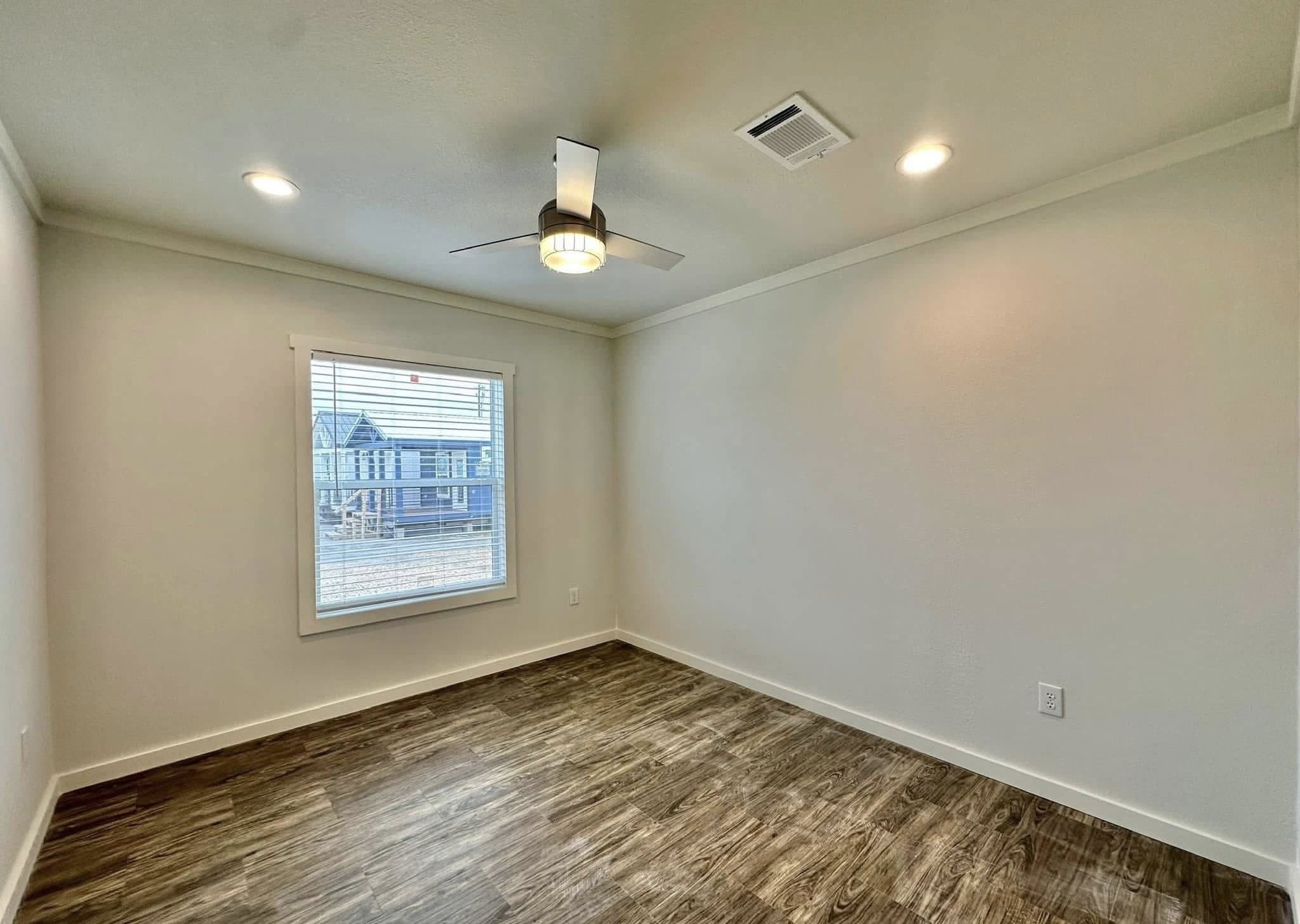 Empty room with wood laminate flooring, white walls, and a large window showing houses outside. A ceiling fan with lights is centrally positioned, giving a modern feel.