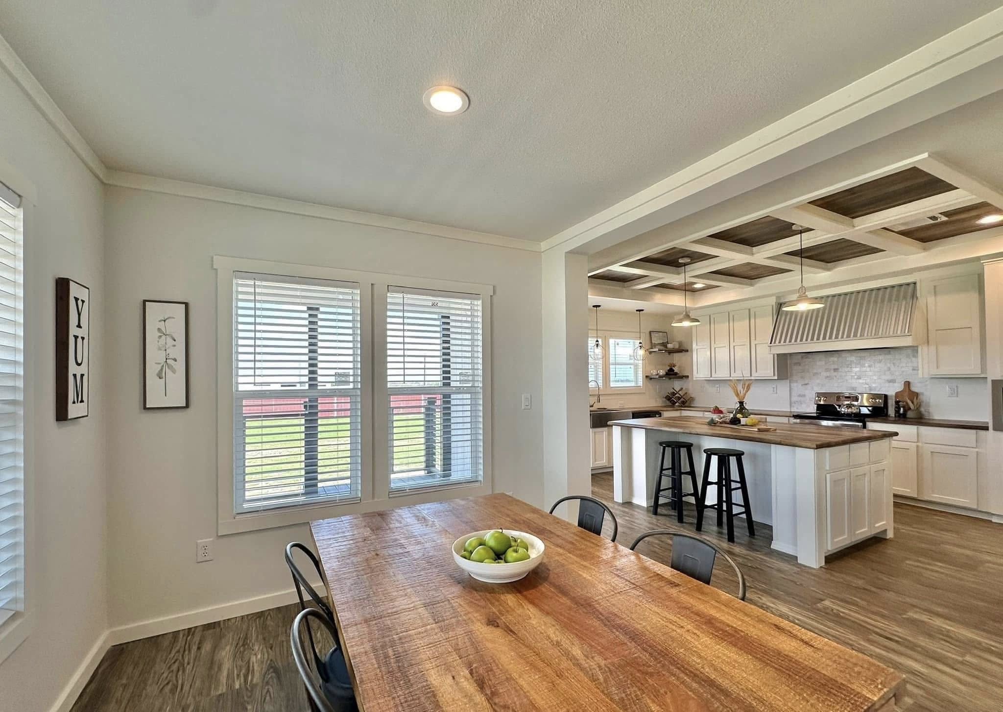 Bright, modern kitchen and dining area with wood floors. A rustic table with a bowl of green apples is in the foreground, while the kitchen features a large island, white cabinets, and pendant lighting.
