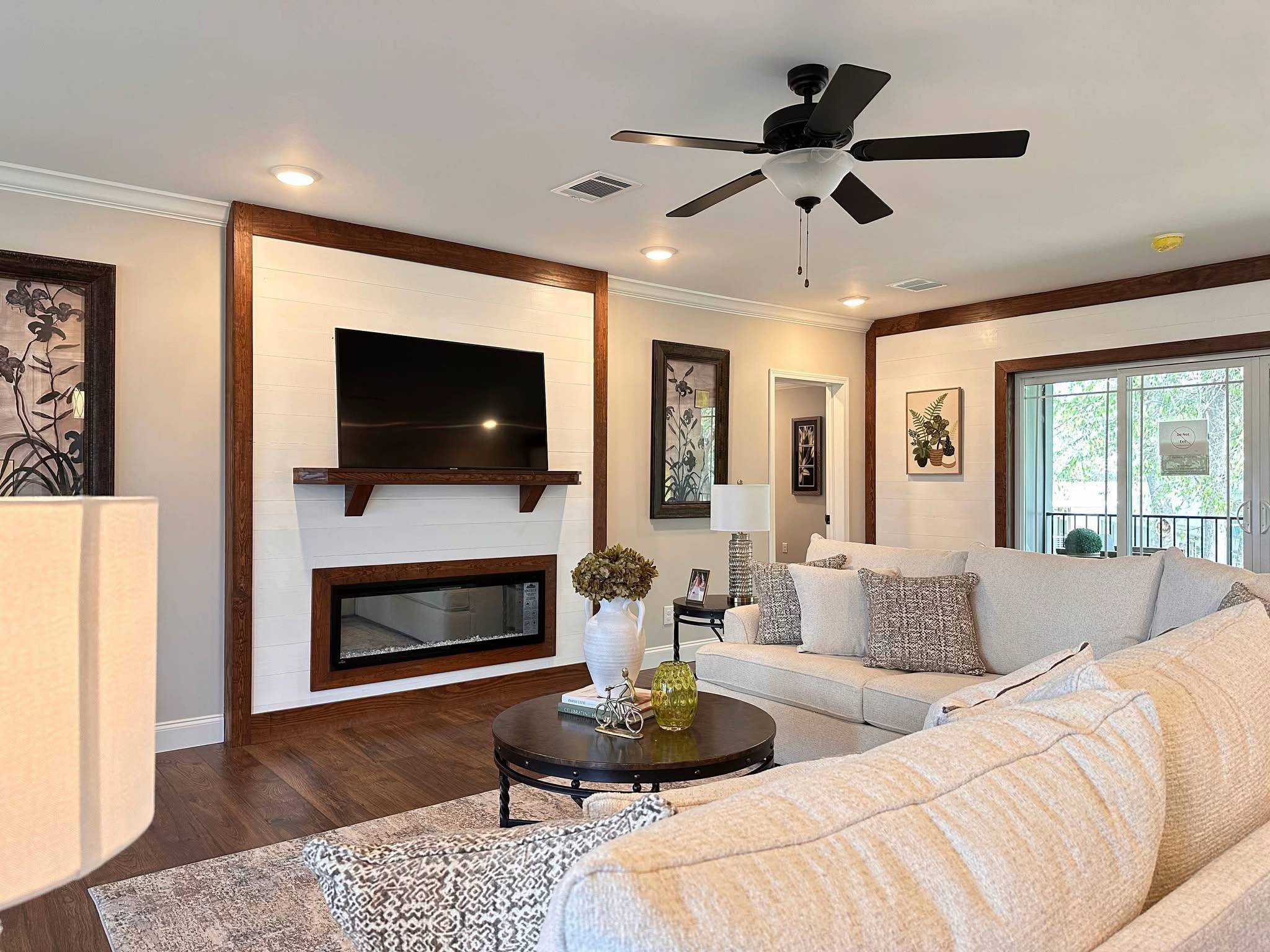 Modern living room with a beige sectional sofa, decorative pillows, and a round coffee table. A wall-mounted TV and fireplace are framed with wood. Natural light filters through large windows, enhancing the cozy, elegant ambiance.