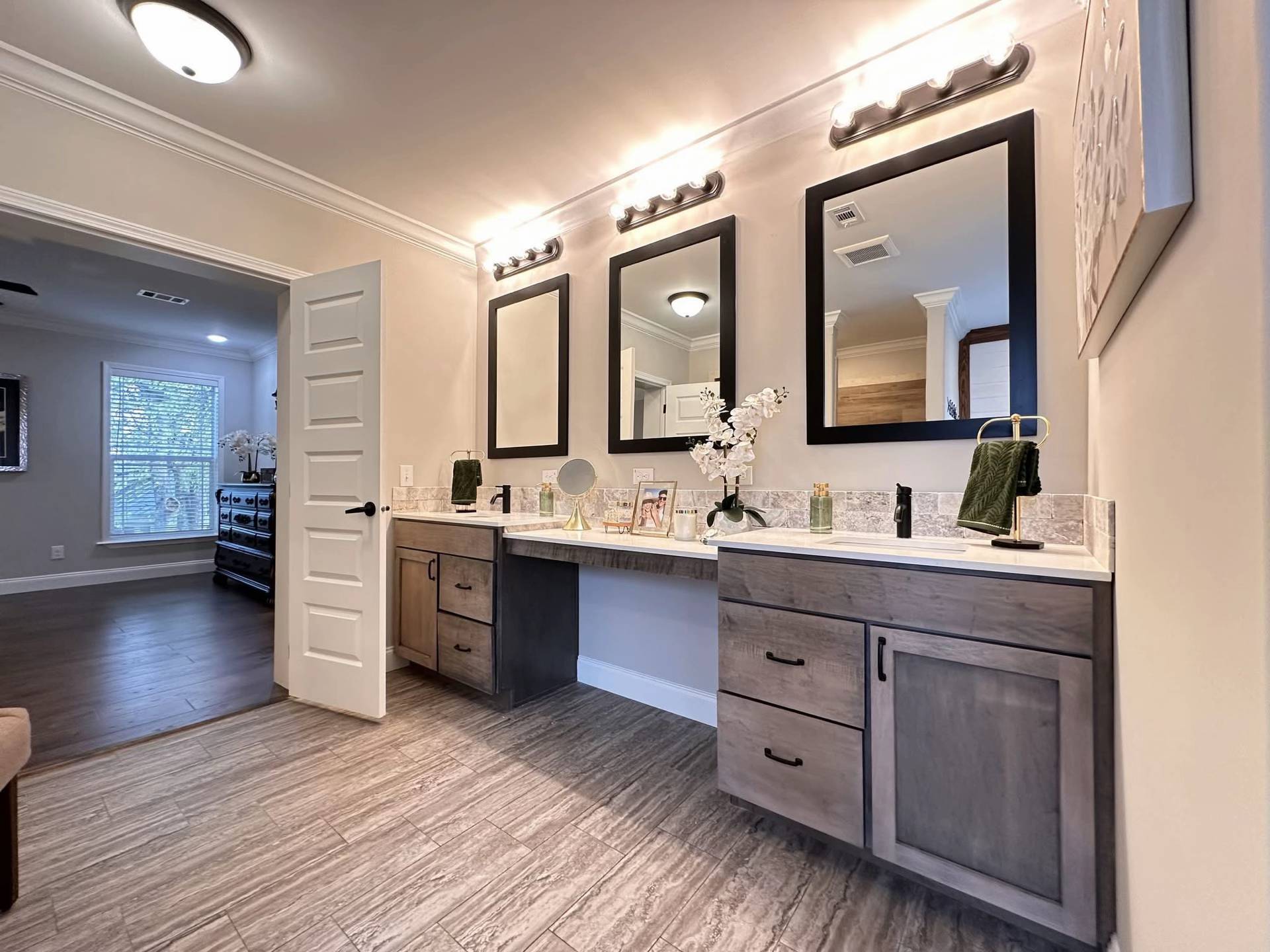 Modern bathroom with wood cabinetry, three framed mirrors, and soft overhead lighting. A doorway leads to a room with hardwood floors, creating a tranquil atmosphere.