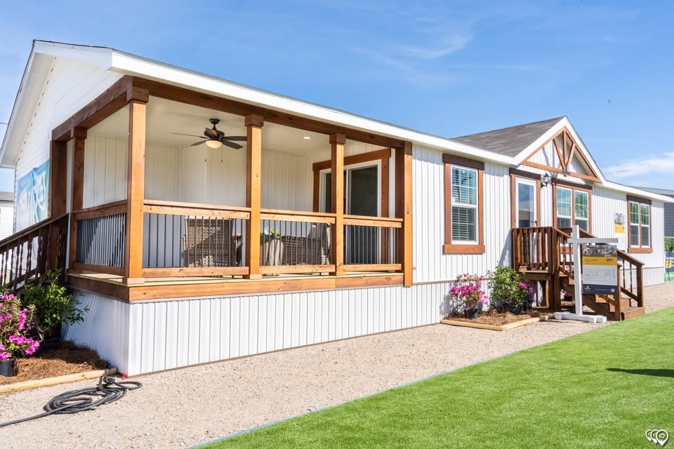 A modern white bungalow with wooden trim and a spacious porch featuring a ceiling fan. Surrounded by green grass and blooming pink flowers under a clear blue sky.