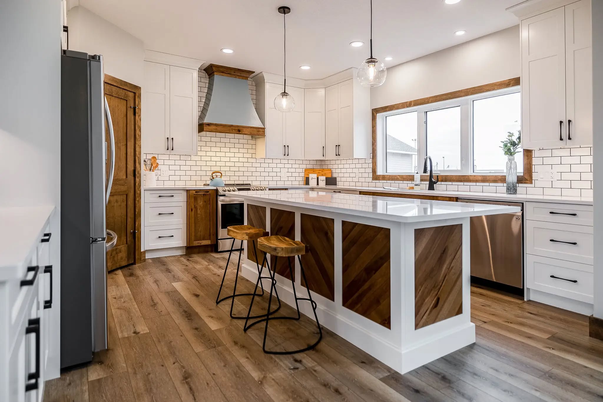 Modern kitchen with a large island featuring wooden accents and two bar stools. White cabinets, stainless steel appliances, and pendant lights create a bright, inviting atmosphere.