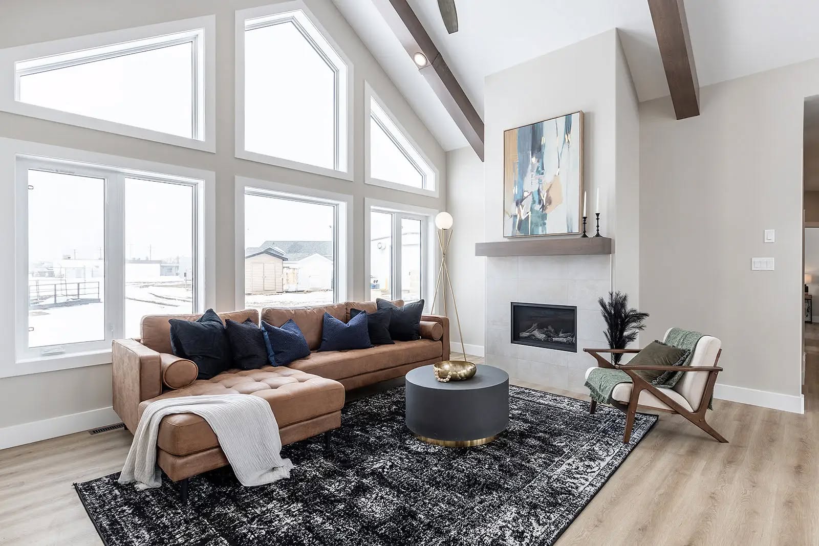 Modern living room with tall windows, light gray walls, and wood beams. Features a tan sofa with blue pillows, a patterned rug, and a cozy fireplace.