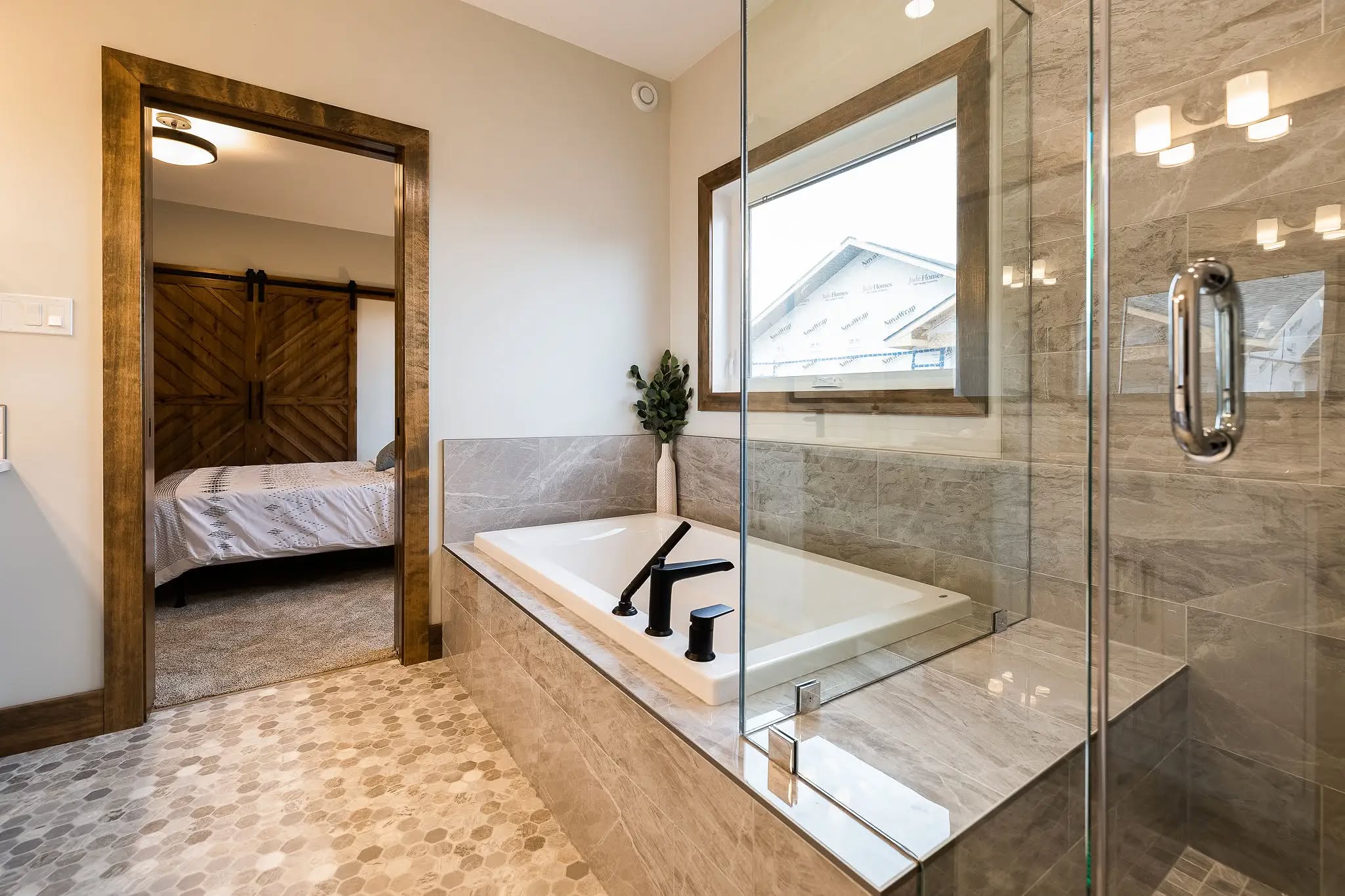 Luxurious bathroom with a glass shower, a large soaking tub, and elegant black fixtures. A barn-style door leads to a cozy bedroom.