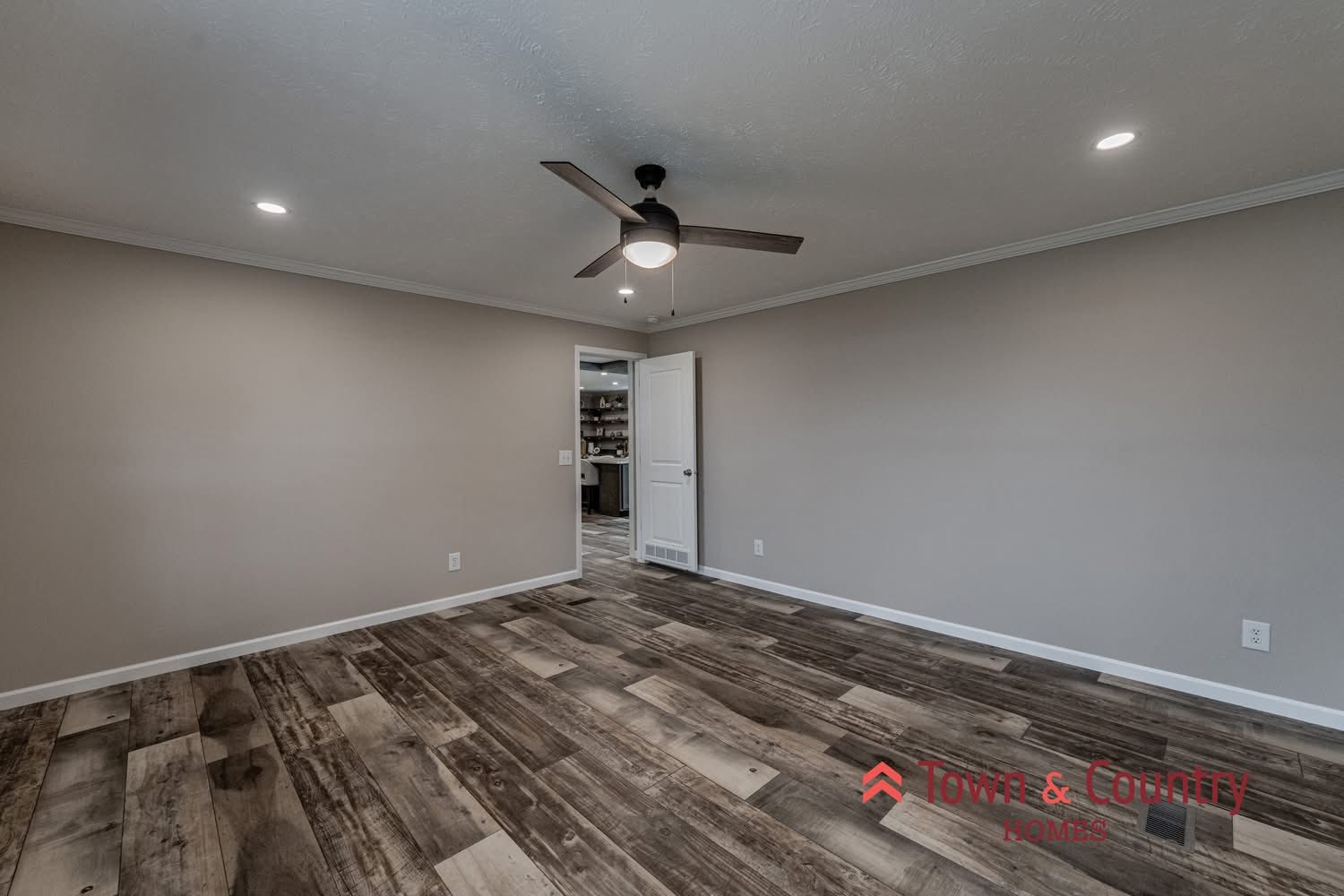 Alt text: "Empty room with wood-look flooring, beige walls, ceiling fan, and recessed lighting. Open door reveals a connecting space, creating a welcoming feel."
