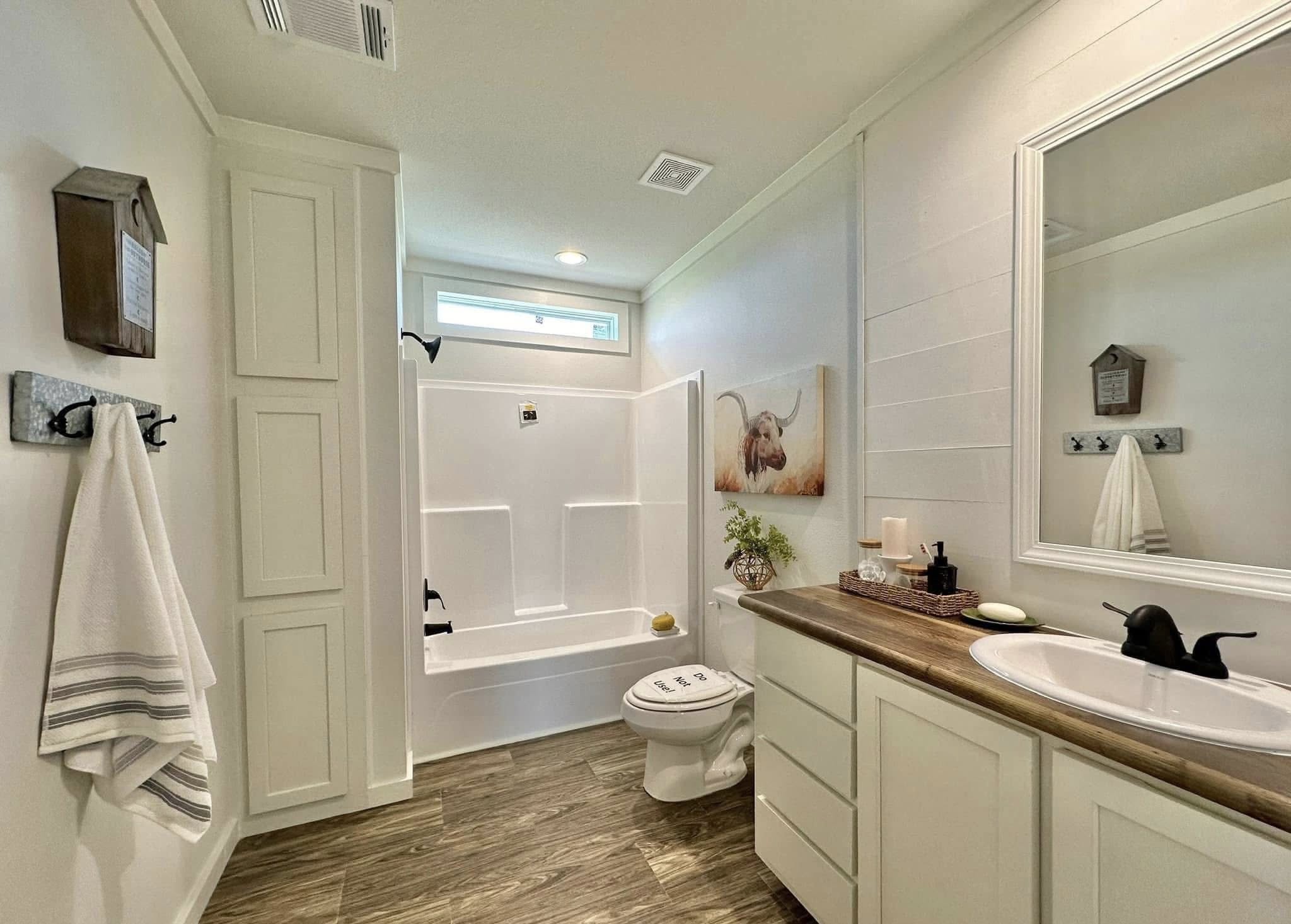 A bright, modern bathroom with wood flooring, white walls, and a shower-tub combo. Features a sink, cabinets, a window, and a decorative wall art.