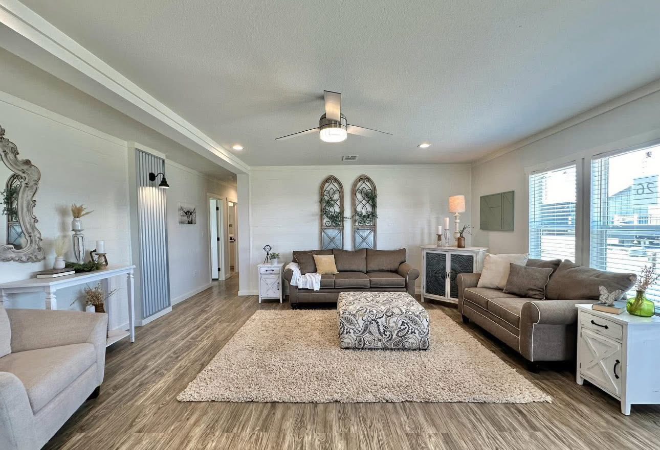Bright living room with two brown sofas and a patterned ottoman on a beige rug. Large windows, elegant decor, and a modern ceiling fan enhance the cozy ambiance.