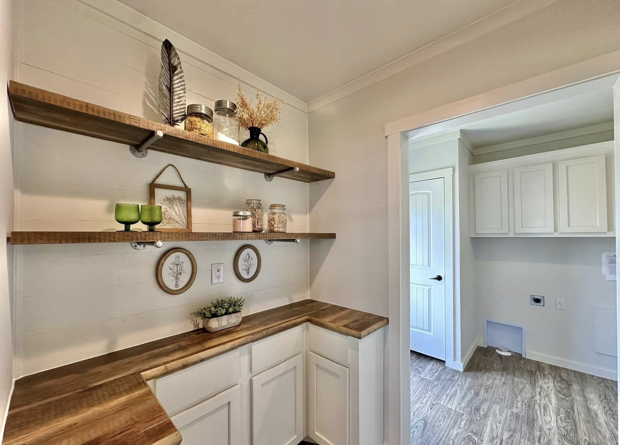 Bright pantry with wooden shelves and white cabinets. Shelves hold jars, a feather, a vase, and plant decor. Adjacent view of laundry room. Cozy and organized.