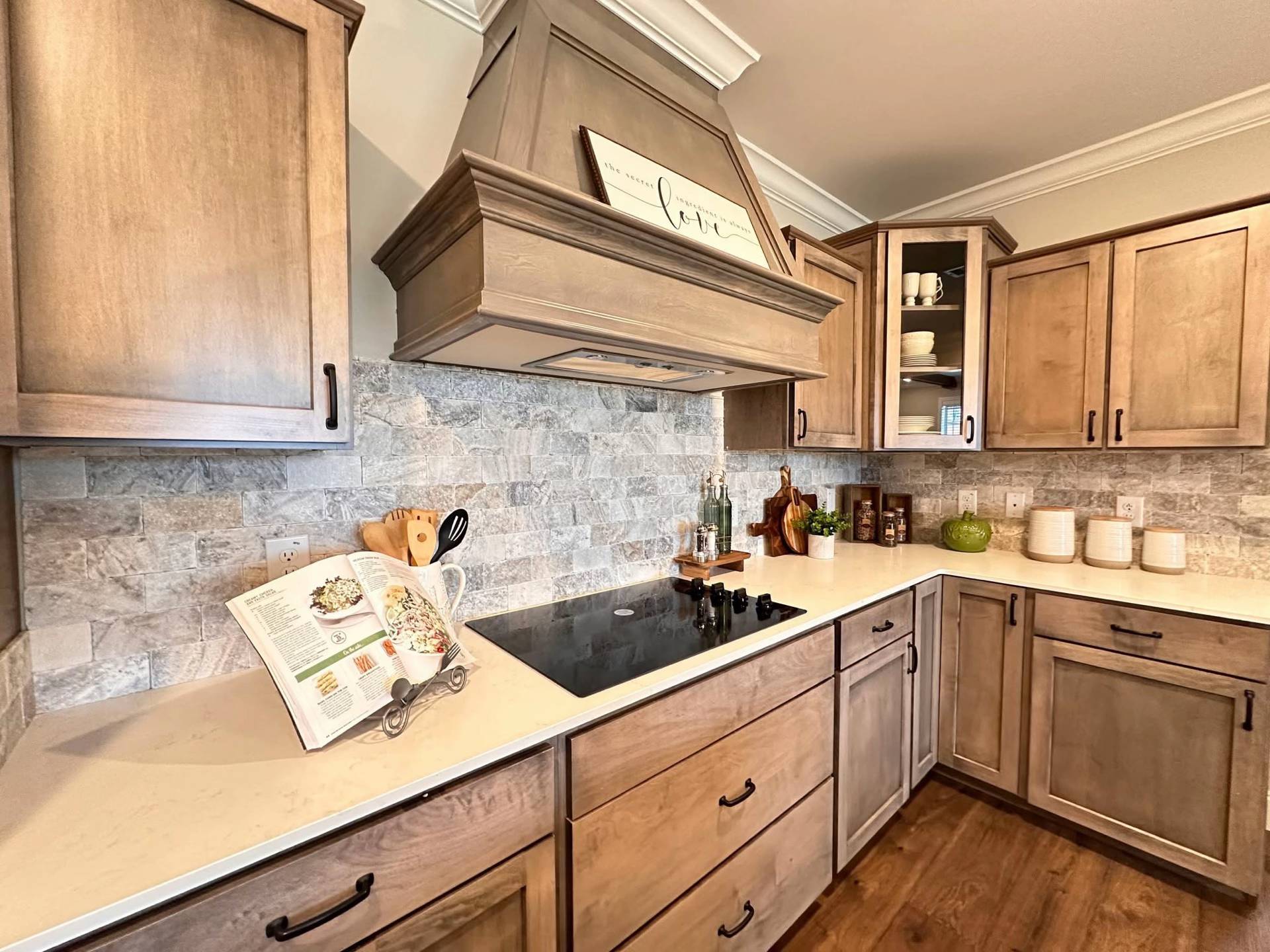 Warm wooden kitchen with a sleek cooktop, cookbook on a stand, rustic decor, and stone backsplash. Cabinets house plates and mugs, creating a cozy feel.