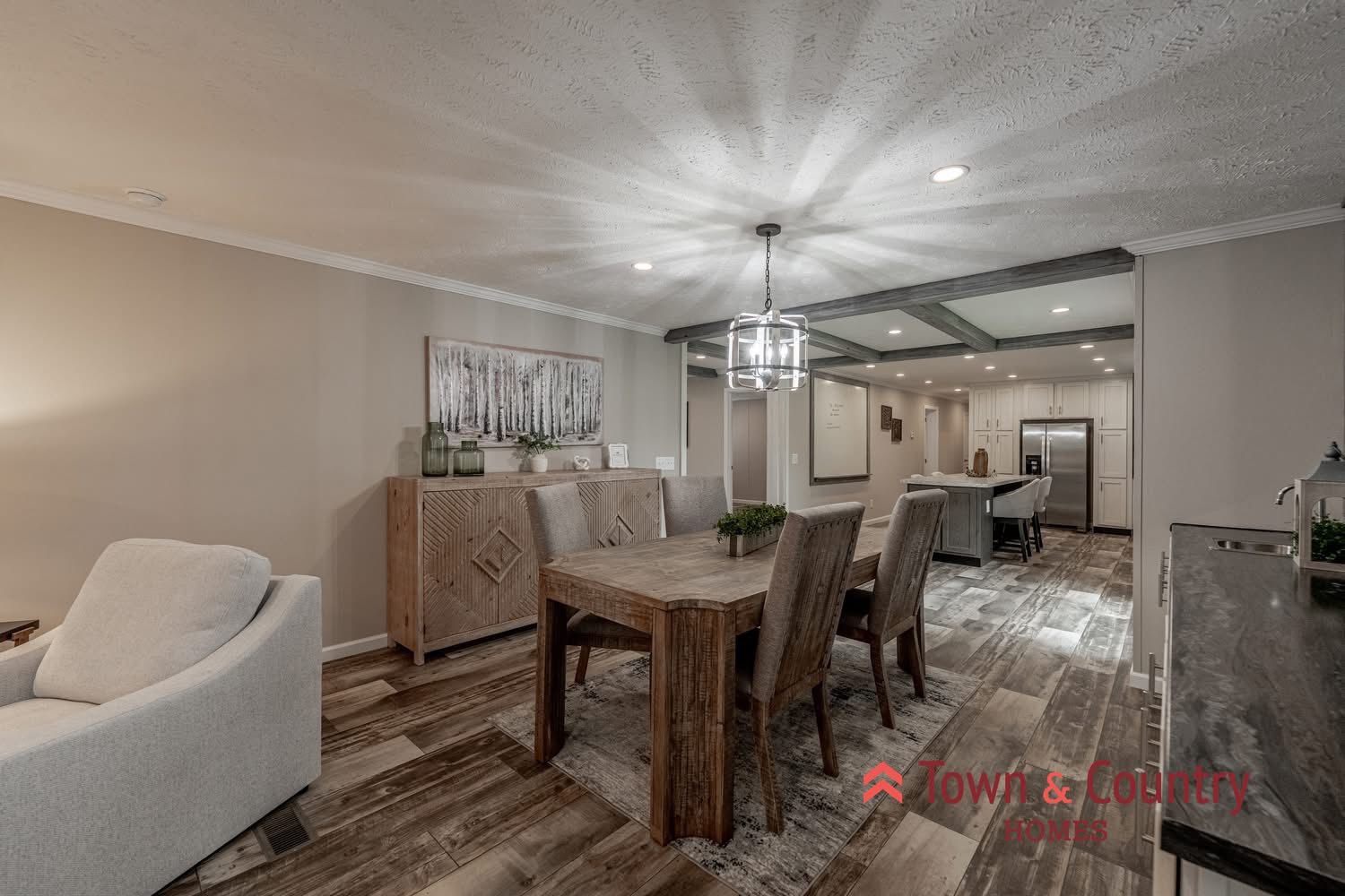 A modern open-concept dining and kitchen area with wooden flooring. A wooden dining table with six chairs is central, under a stylish chandelier. To the left is a light gray armchair. A sideboard with decor and artwork is against the wall. The kitchen in the background showcases white cabinets and stainless-steel appliances. The space feels warm and inviting.