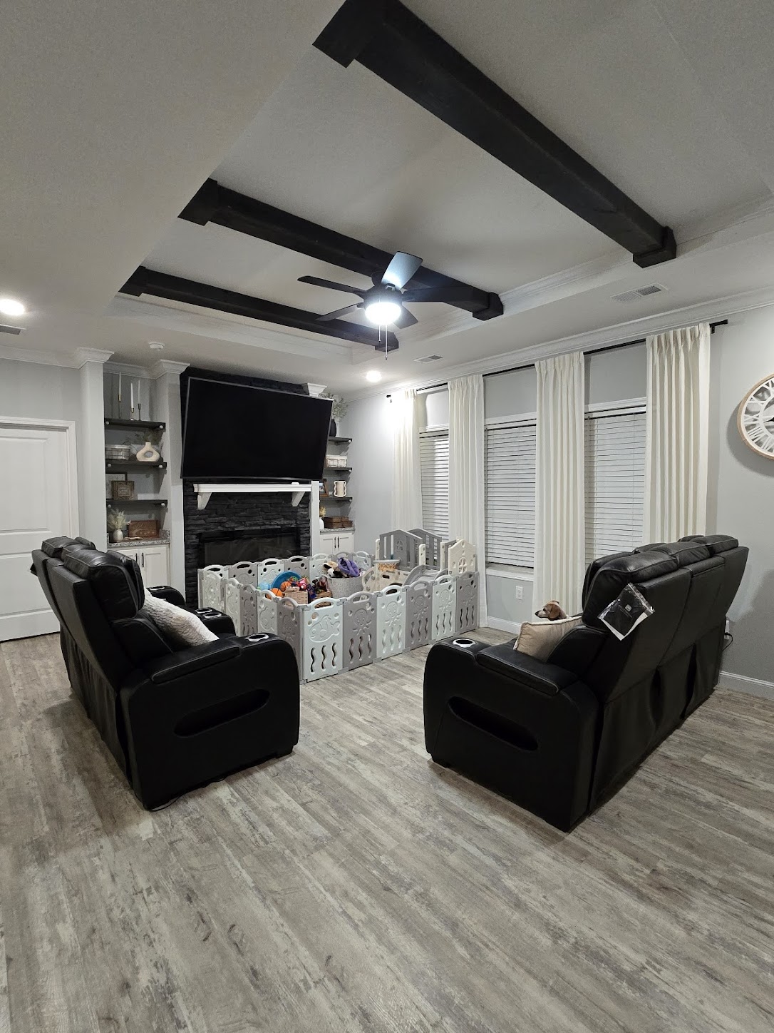 A modern living room features two black recliners facing a fireplace with a flat-screen TV above. A playpen filled with toys is in the center, surrounded by light walls and wooden flooring, creating a cozy atmosphere.