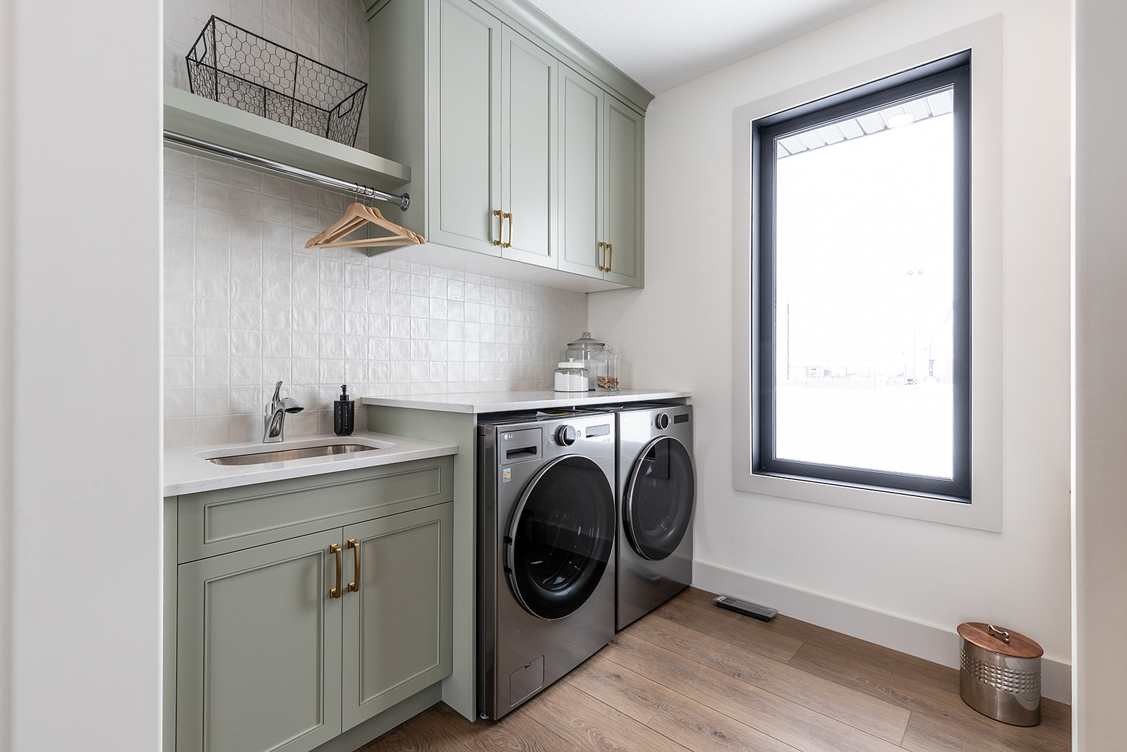 Bright laundry room with green cabinets, light wood floor, and black washer and dryer. Large window on the right, providing natural light.