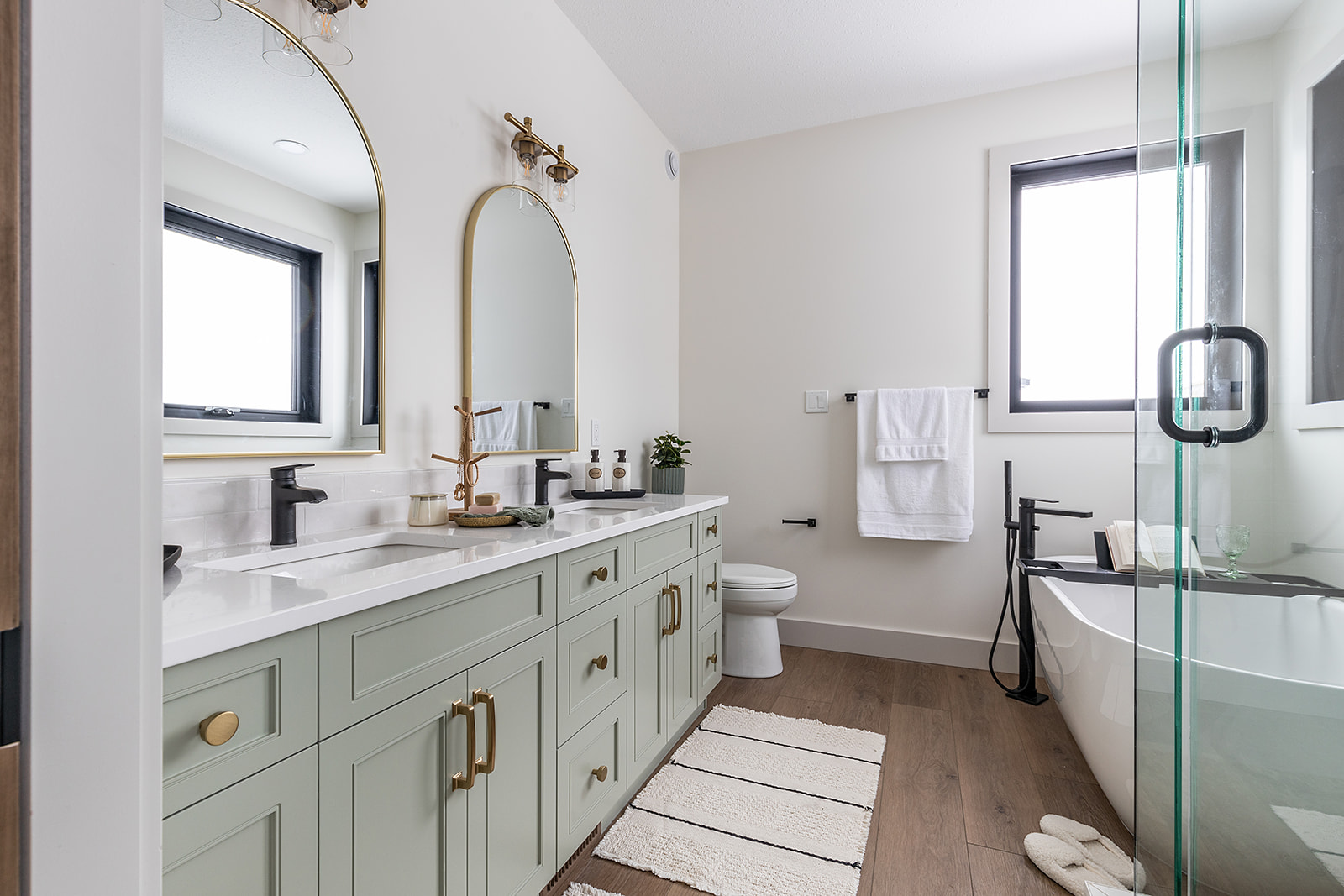 Modern bathroom with mint green double vanity, brass fixtures, and arched mirrors. A freestanding tub is near a window, creating a serene ambiance.
