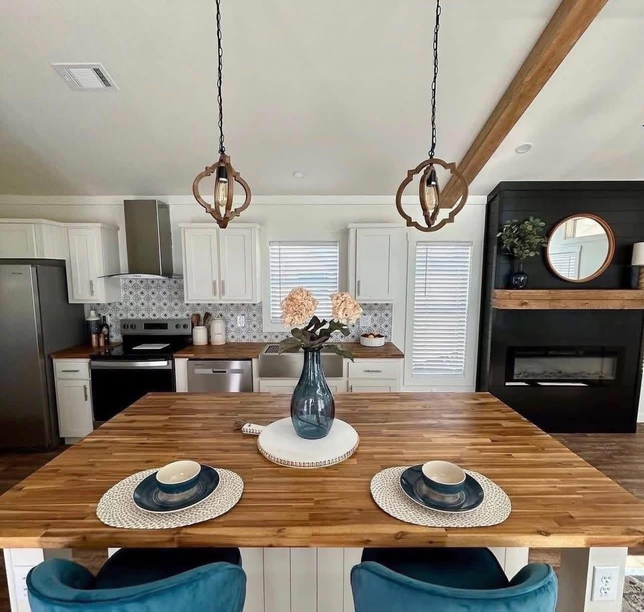 Stylish kitchen with a wooden island, blue chairs, and pendant lights. White cabinets, floral tiles, and a fireplace create a cozy, modern vibe.