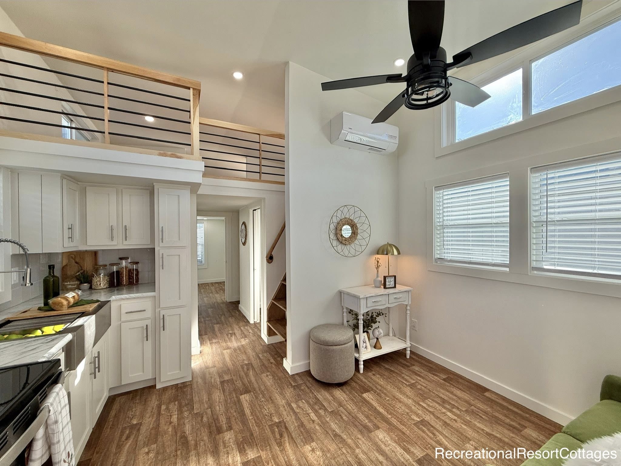 Modern tiny home interior with a loft, light wood flooring, white cabinetry in the kitchen, large windows, a ceiling fan, and cozy decor.