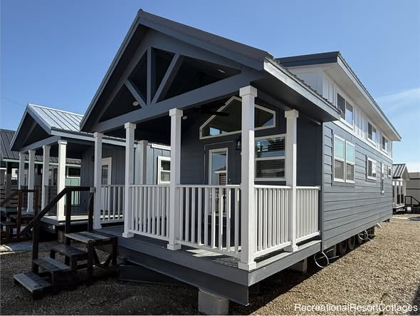 A charming blue tiny house with white trim and a gabled roof. It features a small porch with railings and several windows, under a clear blue sky.