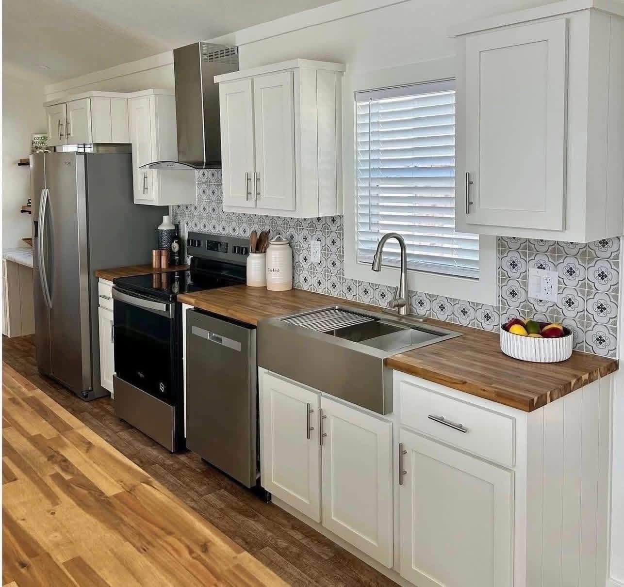 A modern kitchen with white cabinets, stainless steel appliances, and a wooden countertop. A fruit bowl adds color, and a patterned backsplash offers texture.