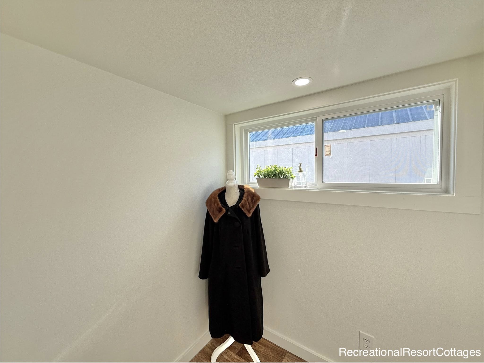 Mannequin with a long, dark coat stands in a bright room with white walls. A small window with a potted plant brings in natural light, creating a serene ambiance.