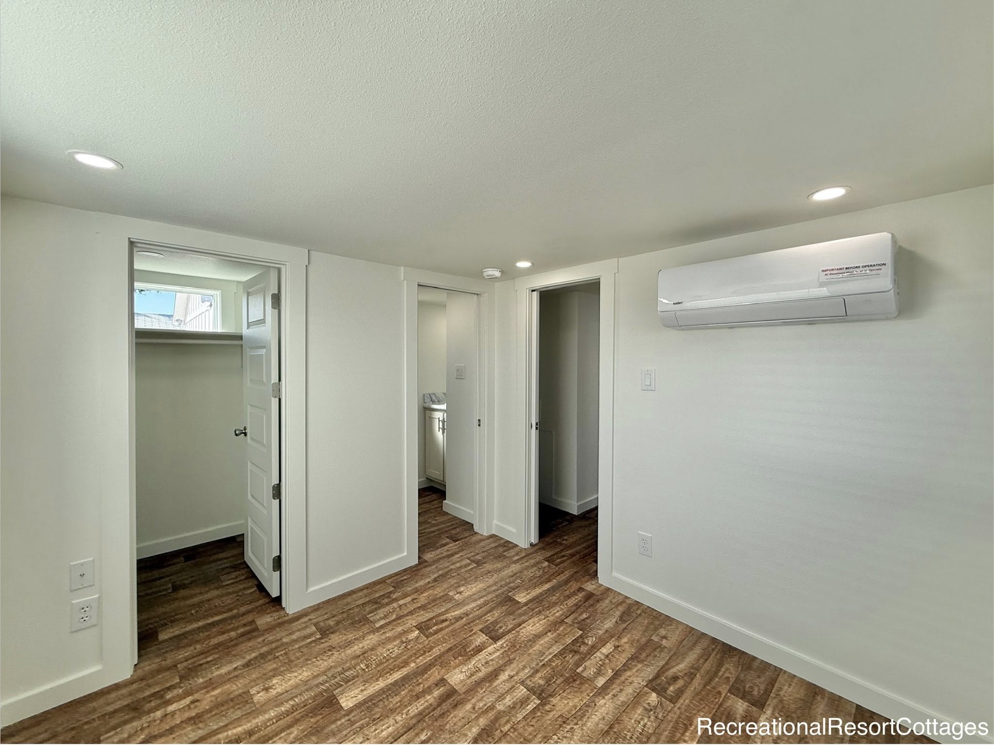 Compact room with wood flooring, two open doorways leading to a closet and bathroom. White walls, ceiling lights, and a wall-mounted air conditioner. Clean and minimal.