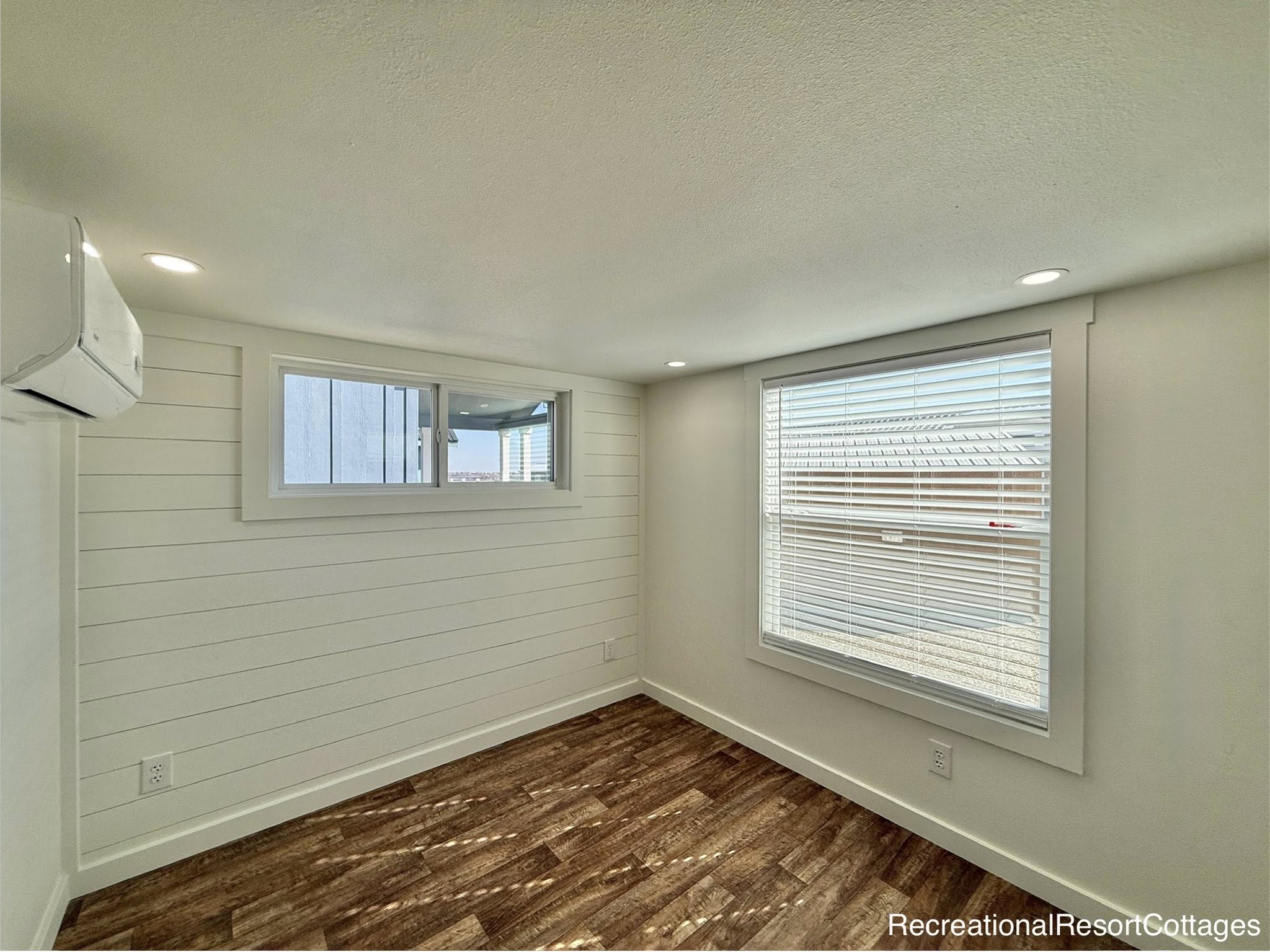 A small, bright room features wooden floors, white shiplap walls, and two windows bringing in natural light. The room has a contemporary, minimalist feel.