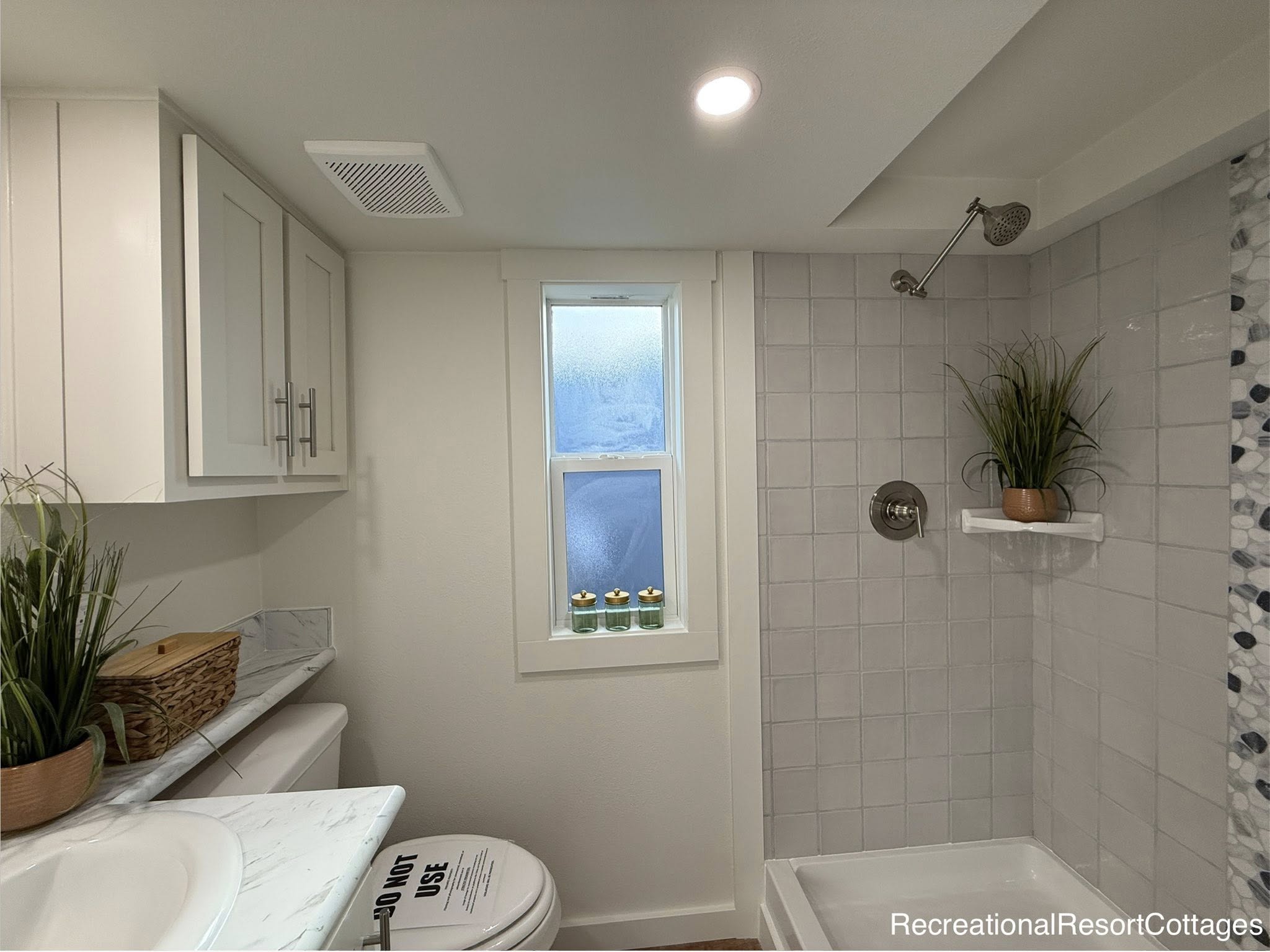 Modern bathroom with light gray tiles, a frosted window, and a small plant on a shower shelf. White cabinets and a marble countertop are visible.