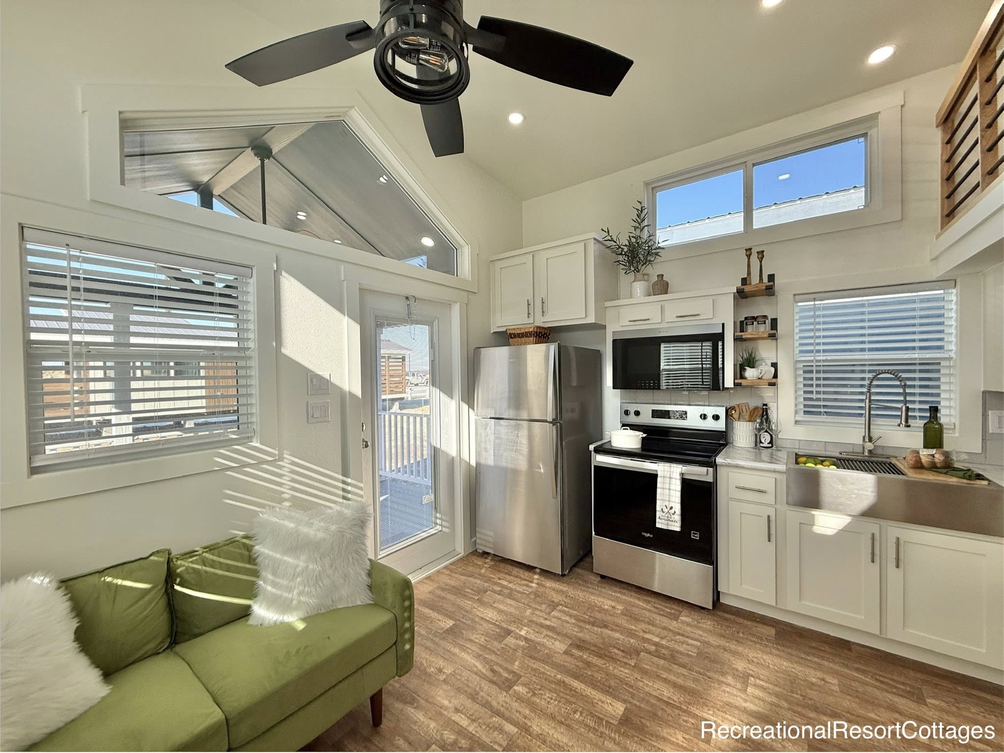 Compact, modern kitchen with white cabinets, stainless steel appliances, and wood floors. A ceiling fan hangs above a cozy green couch, next to a sunlit window.