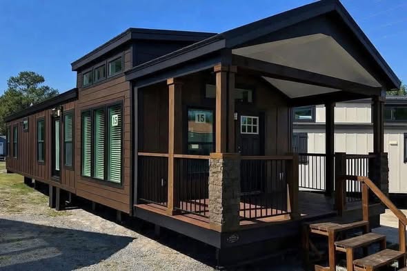 Modern tiny house with dark wood exterior, large windows, and a small porch. The setting is sunny and surrounded by other mobile homes, conveying a cozy feel.