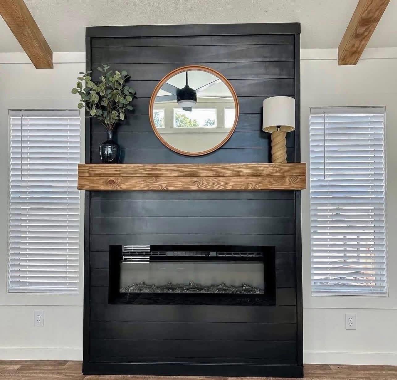 A modern living room features a black shiplap wall with a wooden mantel. A round mirror hangs above, flanked by a potted plant and a twisted rope lamp.