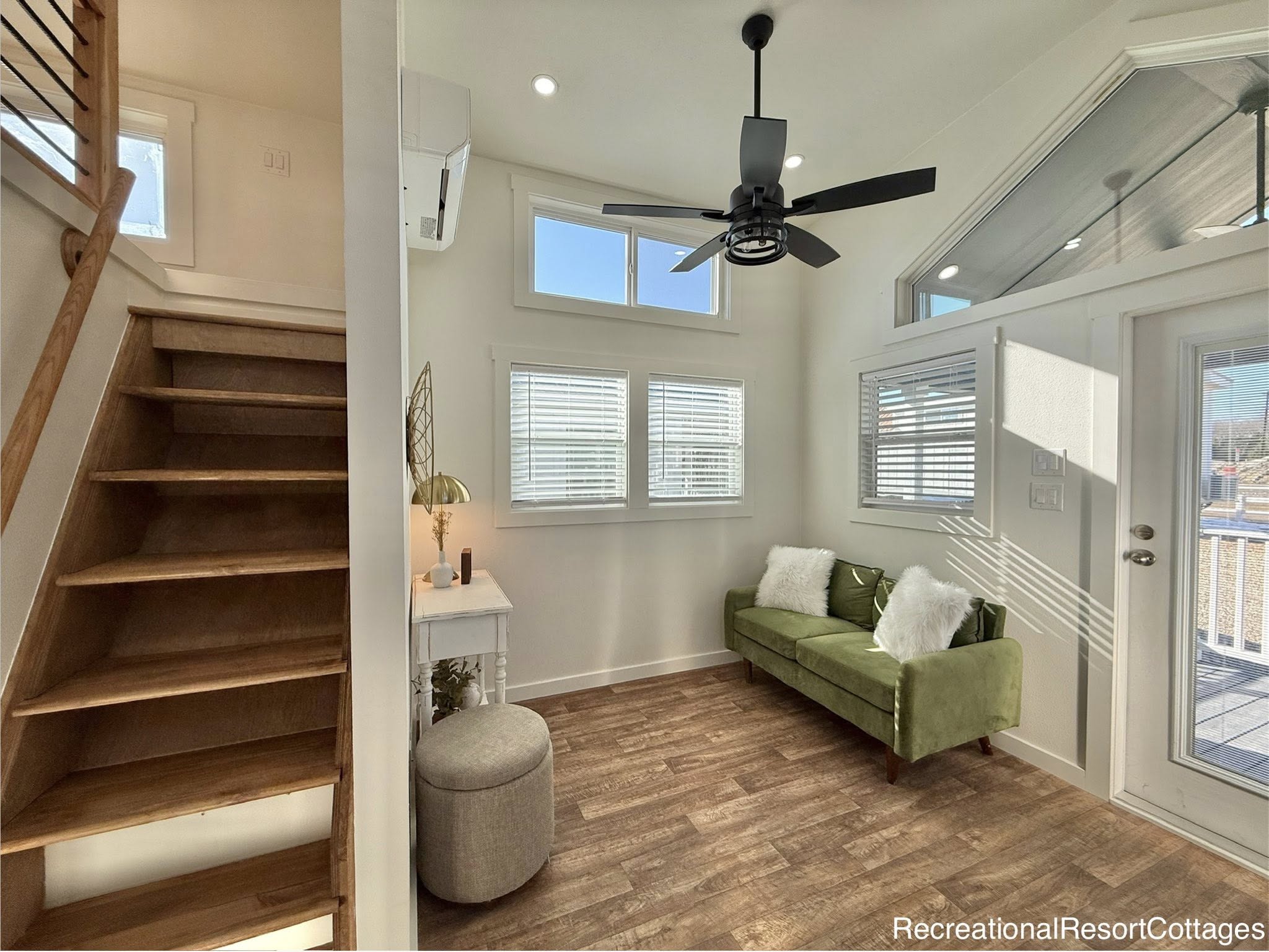A modern, compact living room with a high ceiling, featuring a green sofa and white pillows. Large windows, a ceiling fan, and wooden stairs add charm.