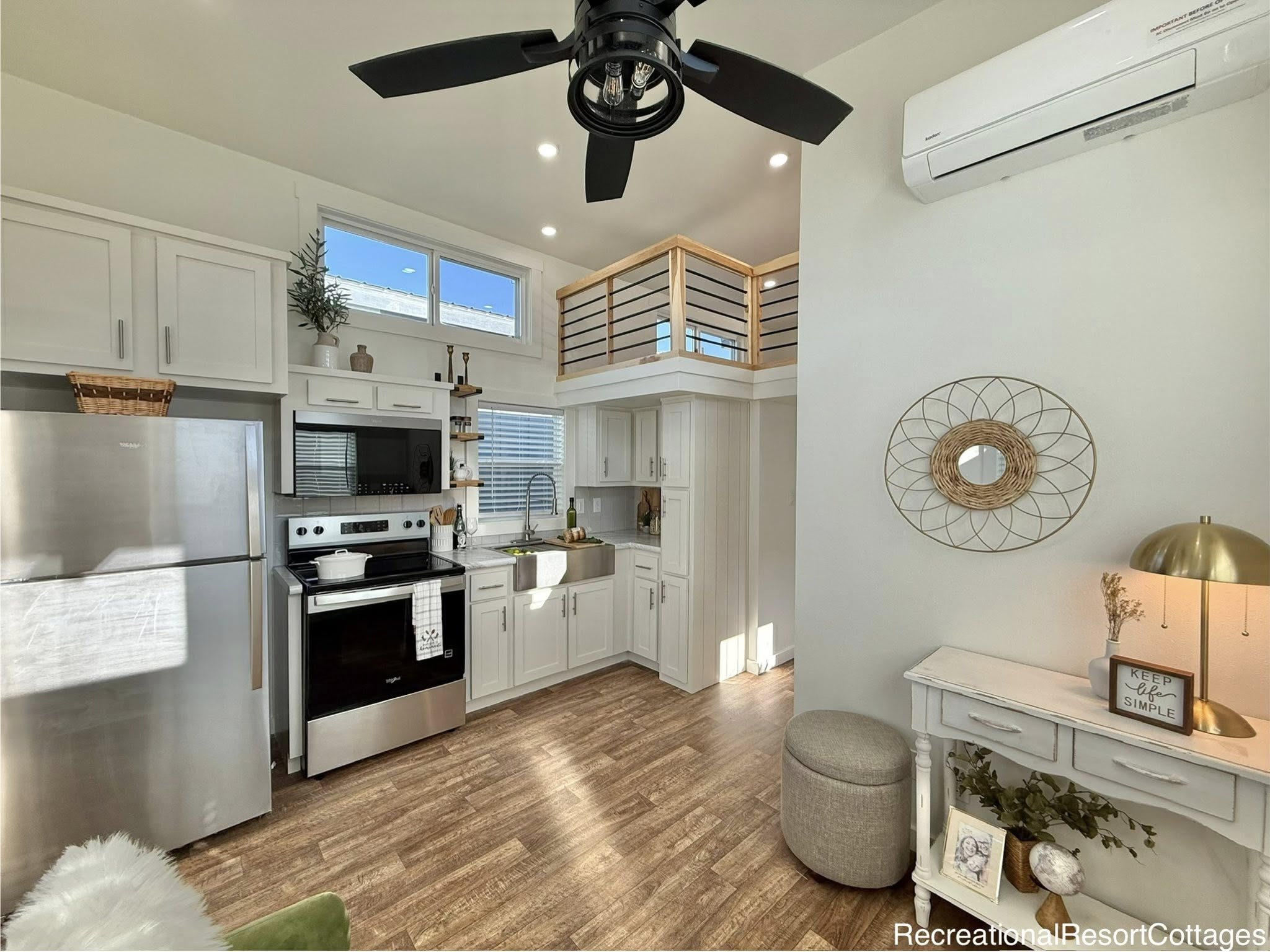Modern tiny home kitchen with stainless steel appliances, white cabinets, and a loft. Warm natural light enhances the cozy, minimalist decor.