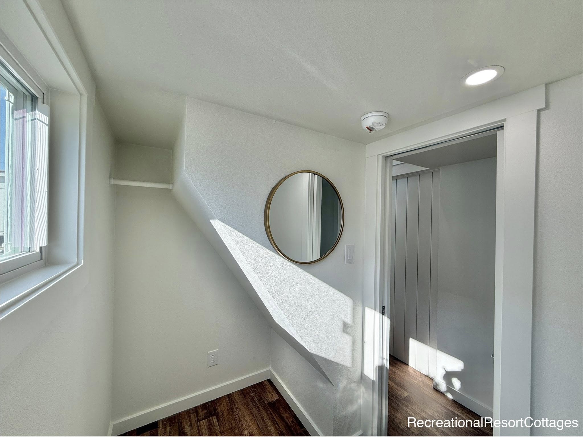 A small, bright room with white walls features a round mirror on one wall and a window on the left. Sunlight casts geometric shadows on the walls. The wood floor adds warmth, conveying a minimalist and cozy atmosphere.