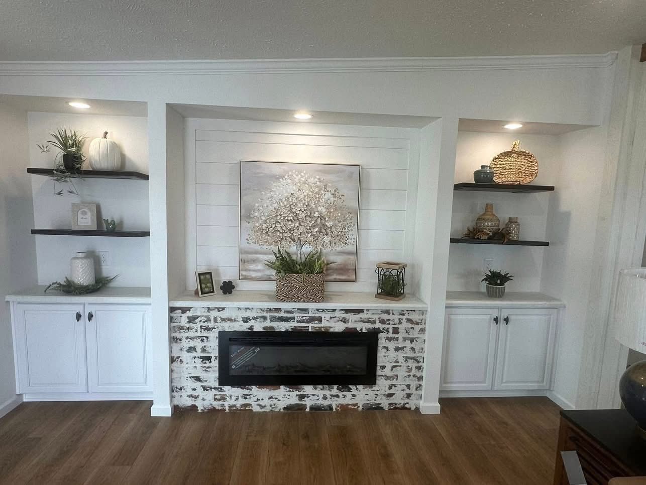 A cozy living room features a white shiplap wall with a floral painting, flanked by built-in shelves displaying decor items. A brick fireplace adds warmth.