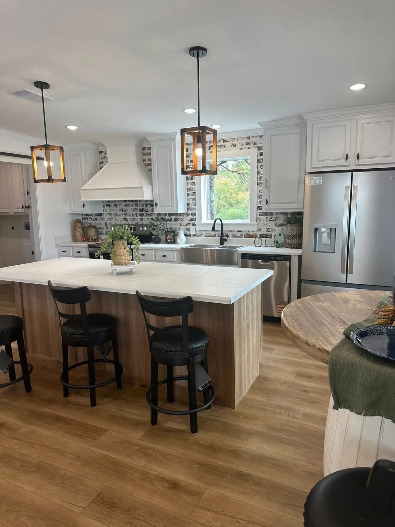 Chic kitchen with a large island, three black stools, pendant lights, stainless steel appliances, and a window above the sink, exuding warmth.