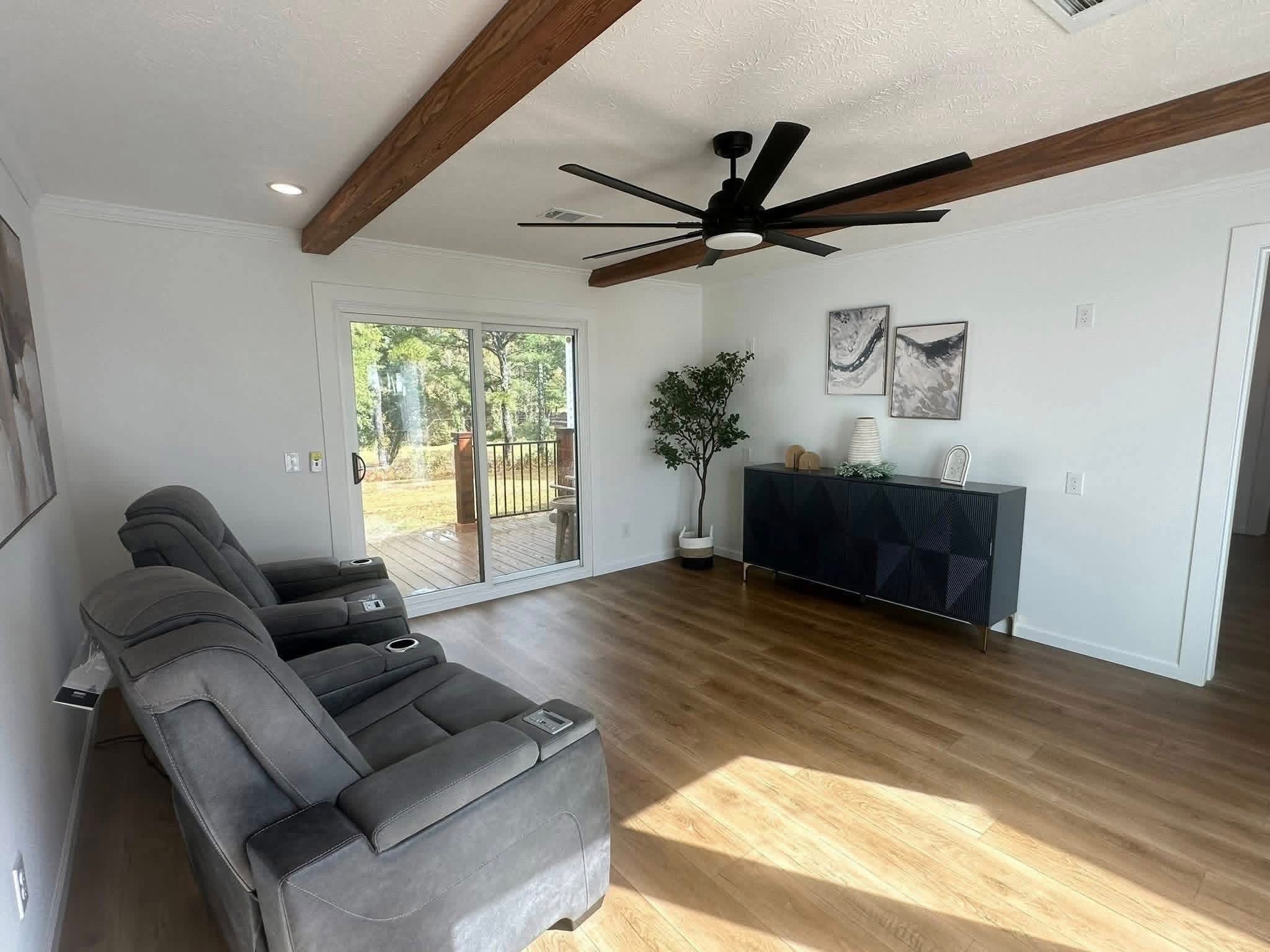 Modern living room with wooden floors, two gray recliners, a large ceiling fan, and sliding glass doors opening to a deck. Cozy, minimalist decor.
